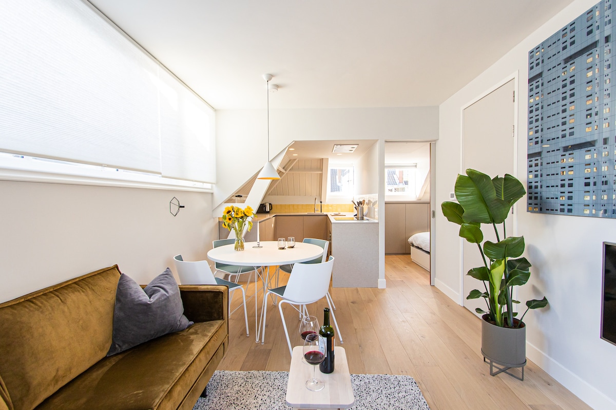 A modern living space features a plush brown sofa and a small table set for two. The bright kitchen is visible in the background, along with light streaming through large blinds. A green plant adds a natural element, while a cozy rug lies beneath the seating area.