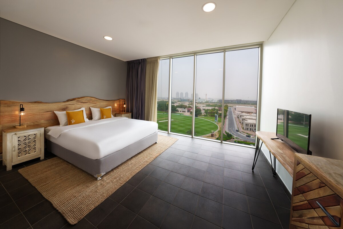 A spacious en-suite bedroom features a comfortable bed with a natural wood headboard. Large floor-to-ceiling windows provide views of the surrounding area. A small television stands on a wooden table, and a woven rug lies on the tile floor.