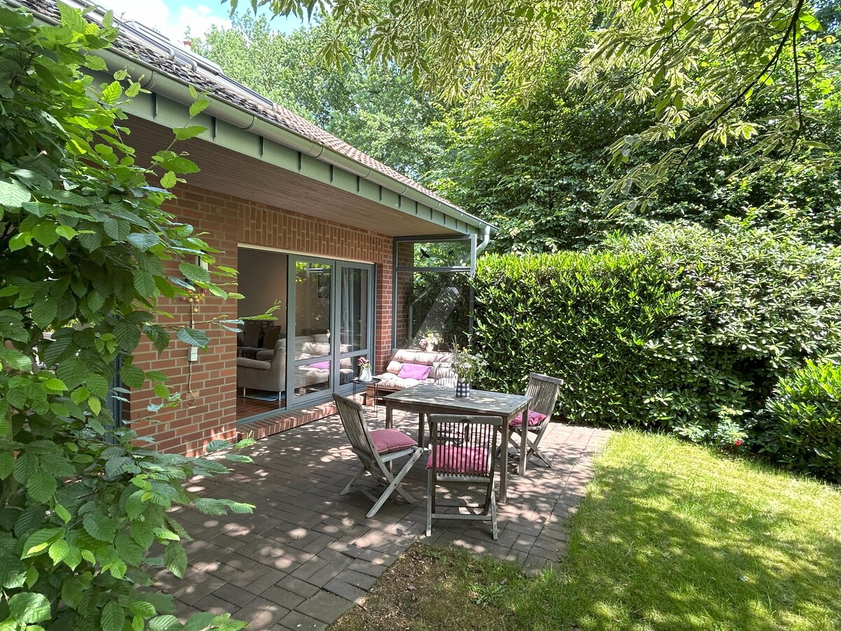 A spacious outdoor terrace is seen surrounded by lush greenery and hedges. Wooden furniture, including a rectangular table and chairs, is arranged for dining. The entrance to the apartment is visible through large glass doors, inviting natural light into the interior.
