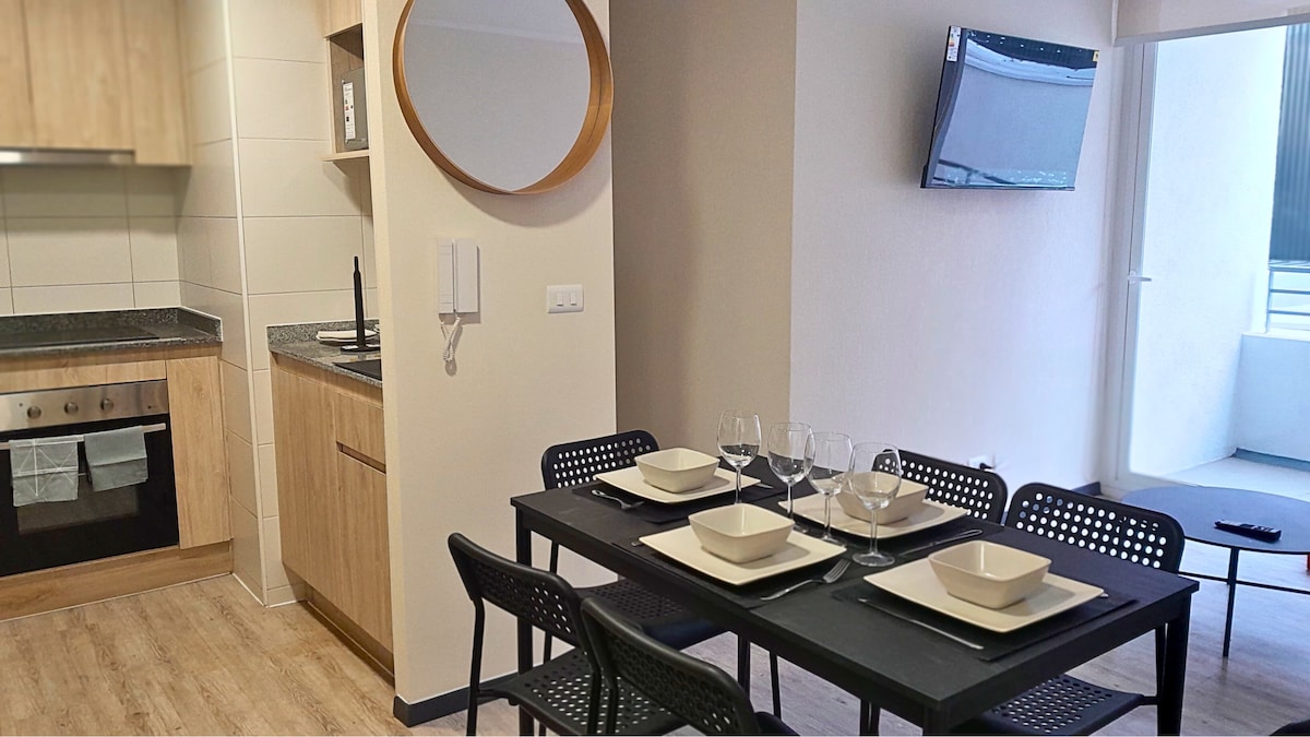 A modern dining area is highlighted by a sleek black table set with plates and wine glasses. A fully equipped kitchen is visible in the background, featuring wooden cabinetry and a stainless-steel oven. Natural light enters through a nearby window, complementing the casual dining space.
