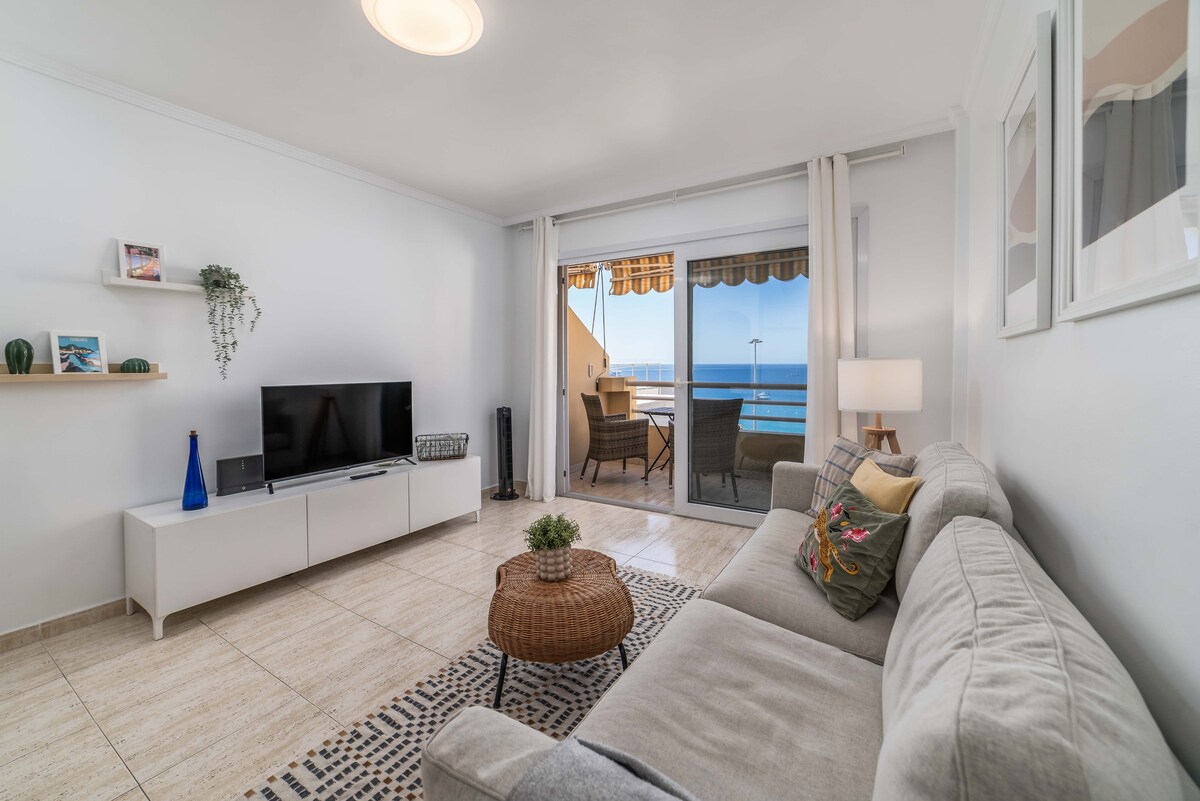 A comfortable living area is presented, featuring a light-colored sofa and a decorative coffee table. A flat-screen television is mounted on a white media console against the wall. Natural light filters in through sliding glass doors that open to a balcony with ocean views.