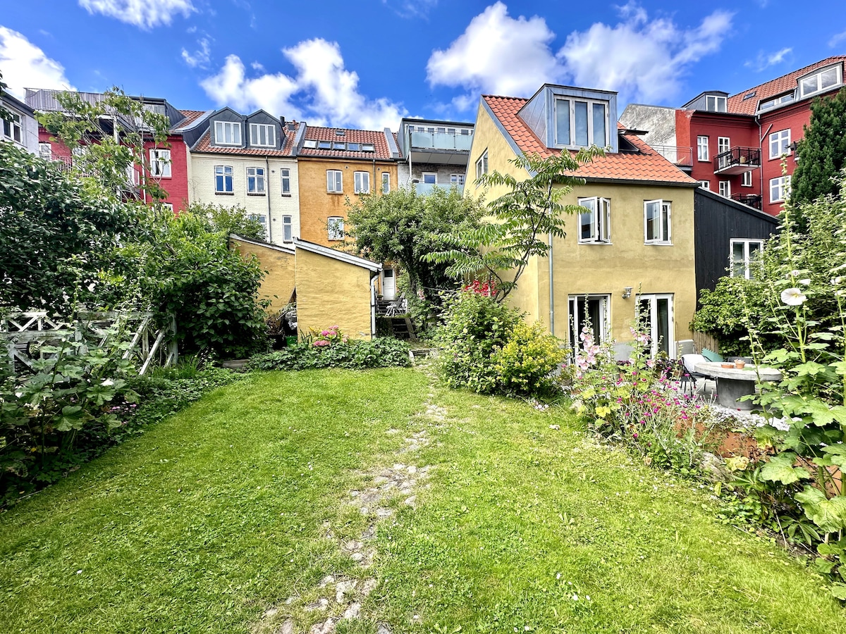 A green garden space is framed by vibrant multi-colored buildings. A grassy area is complemented by various plants and flowers, leading to a yellow house with large windows. A mix of residential architecture creates a charming urban backdrop against a partly cloudy blue sky.