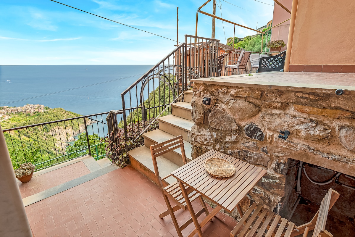 A private terrace with a wooden table and chairs is visible, surrounded by lush greenery. Steps lead down from the terrace, offering an unobstructed view of the sea and nearby coastal landscape, enhancing the outdoor experience.