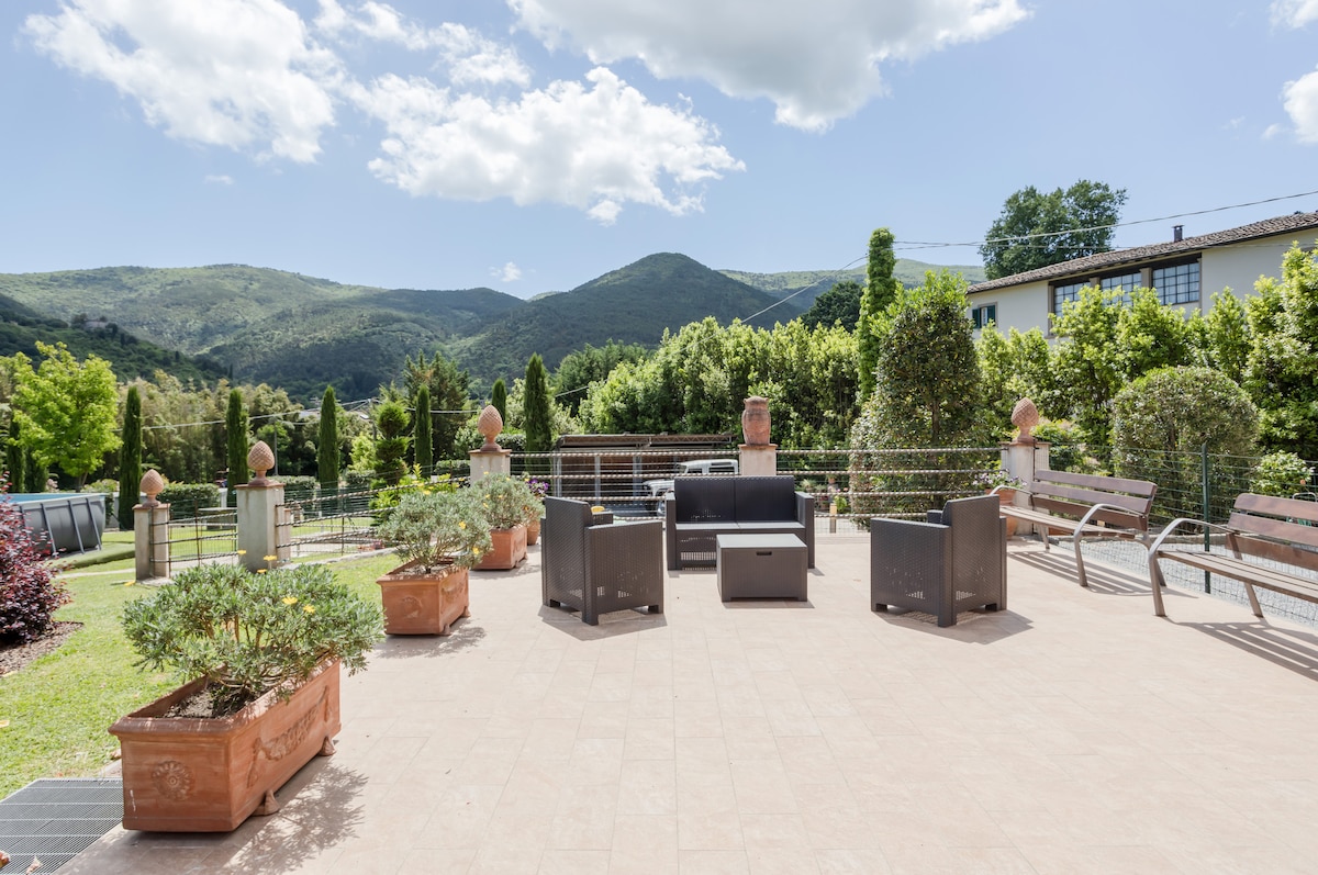 An outdoor patio area is depicted, featuring several seating arrangements with modern chairs. Lush greenery and mountainous scenery are visible in the background, providing a tranquil atmosphere. The space is highlighted by terracotta planters filled with various plants, creating an inviting setting.