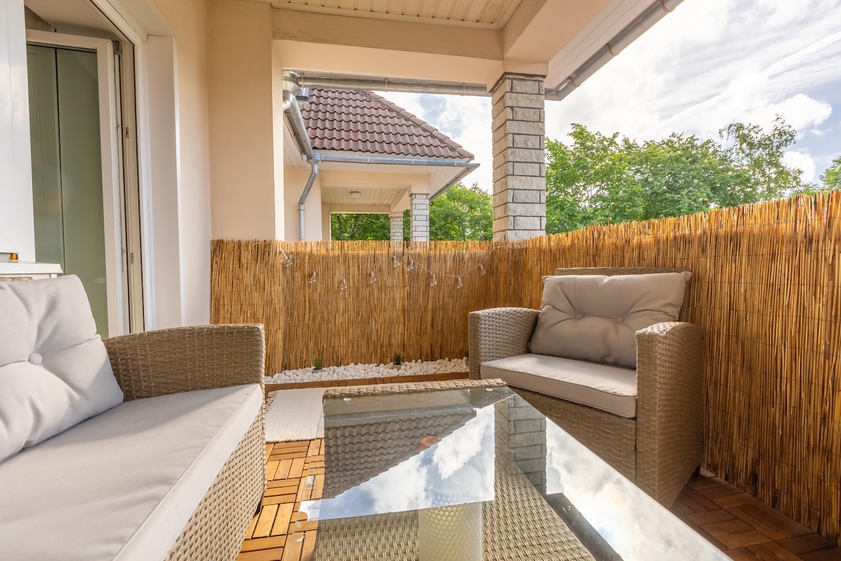 A cozy balcony is furnished with two comfortable seating chairs and a glass-topped table. Natural light brightens the space, and greenery is visible in the background. A bamboo privacy screen adds a sense of seclusion, contributing to a tranquil atmosphere.
