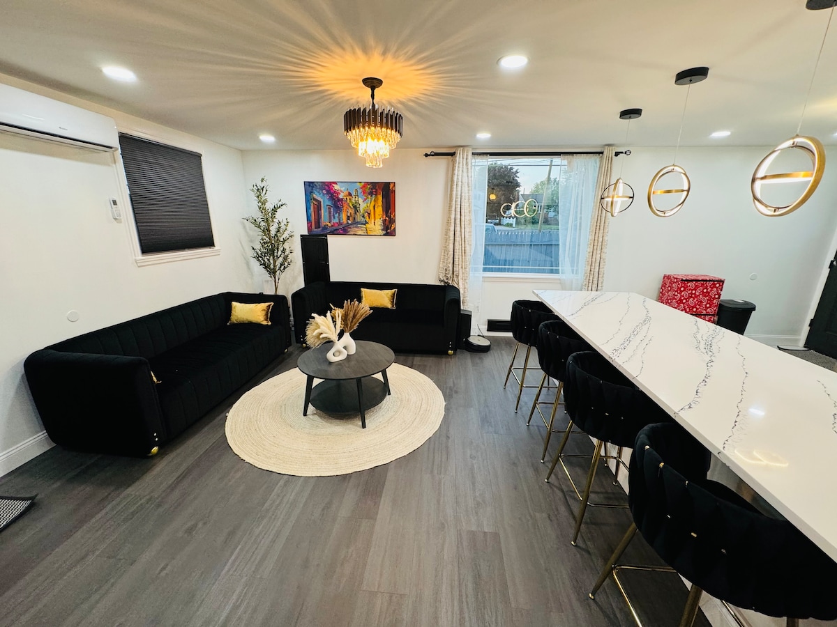 An open-concept living area features a black velvet sofa with gold cushions, complemented by a round coffee table atop a light area rug. A modern dining space is highlighted by a white marble countertop and sleek black barstools, illuminated by stylish pendant lights.