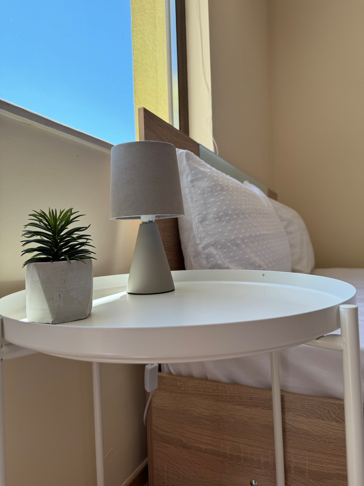 A bedside table holds a small potted plant and a modern lamp. A spacious bed is adorned with white linens, while sunlight spills through a nearby window, creating a bright and airy feel.