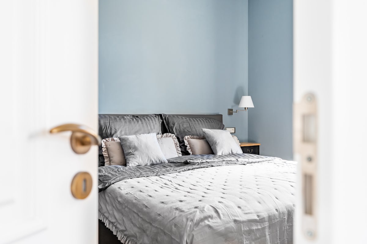 A serene bedroom is visible through an open door. A spacious bed dressed in soft grey linens is complemented by decorative pillows. A bedside lamp casts gentle light, while the calming blue walls add to the peaceful ambiance of the room.