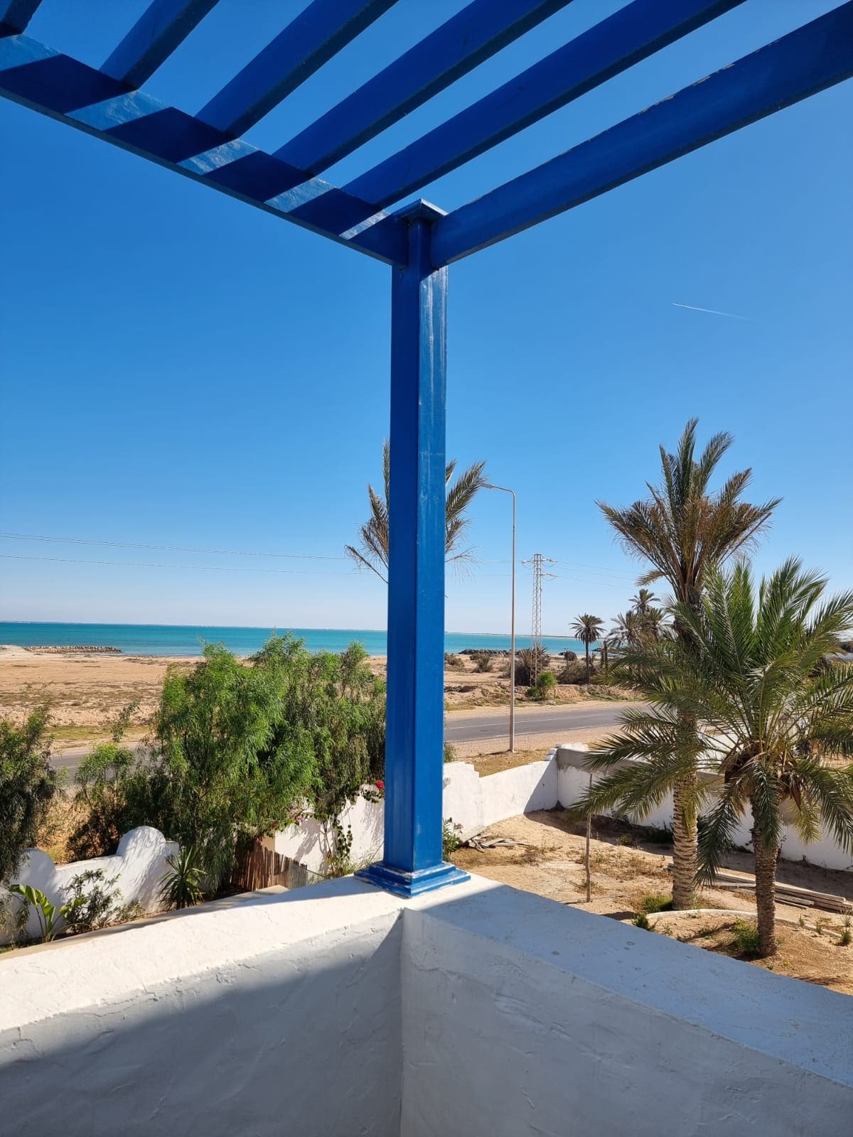 A scenic view from a balcony is presented, showcasing a sandy beach and the calm sea beyond. Tall palm trees frame the coastline, while a clear blue sky enhances the peaceful atmosphere. The balcony's blue railing adds a pop of color to the natural surroundings.