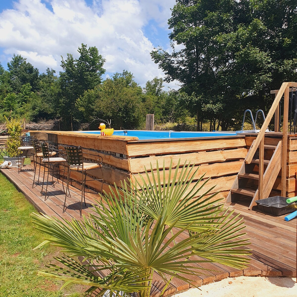 A charming outdoor pool is set within a wooden deck, surrounded by lush greenery. Barstool seating lines the edge of the deck, and a small palm plant adds a touch of nature. Sunshine highlights the inviting blue water, enhancing the overall serene atmosphere.