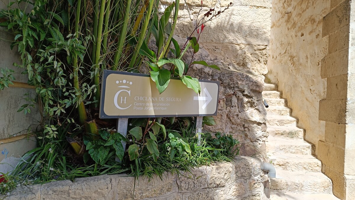A sign indicating the direction to the local site is surrounded by lush greenery. The sign features a prominent logo and text, pointing right towards a pathway with stone steps leading upward, framed by natural elements.