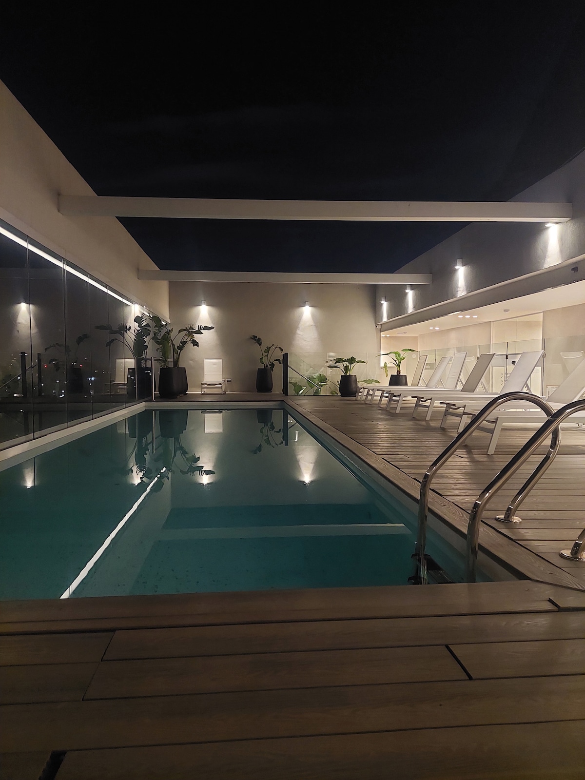 An indoor pool area is illuminated by soft lighting, surrounded by potted plants and sleek lounge chairs. The clear water of the pool reflects the ambient light, creating a serene atmosphere. Glass walls provide views of the exterior while maintaining a private environment.