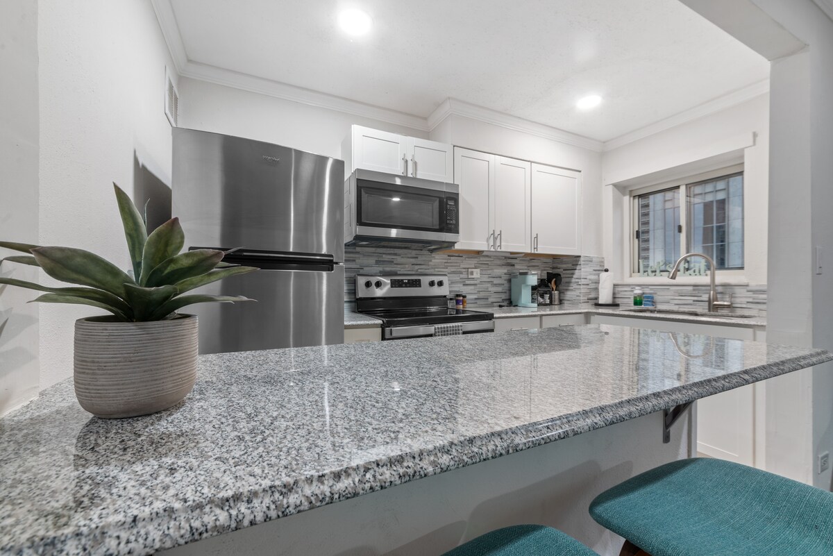The kitchen is equipped with modern stainless-steel appliances and sleek granite countertops. A well-stocked space includes a microwave and coffee maker, with light streaming in from a nearby window. Two turquoise stools provide seating at the breakfast bar, enhancing the functional layout.