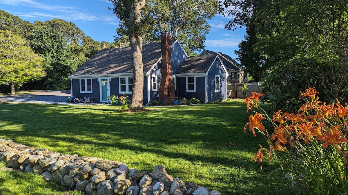 A charming ranch-style home is presented, surrounded by a spacious green lawn and meticulously landscaped flower beds. A stone-lined path leads to the entrance, where vibrant orange flowers add a touch of color. The exterior features a combination of siding and a brick chimney.