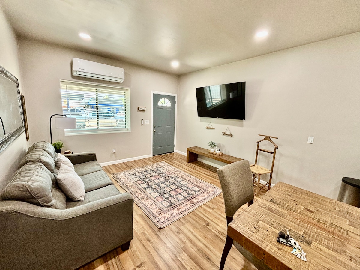 A modern living area features a comfortable gray sofa facing a wall-mounted television. Natural light enters through the window adorned with blinds. A wooden coffee table and a simple dining table with chairs complement the warm wooden flooring.