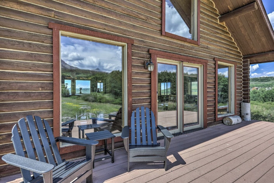 A wooden deck features two Adirondack chairs and a small table, inviting relaxation. Large windows provide expansive views of the surrounding landscape, reflecting the natural scenery and sky. The cabin's log exterior enhances the rustic charm of the setting.