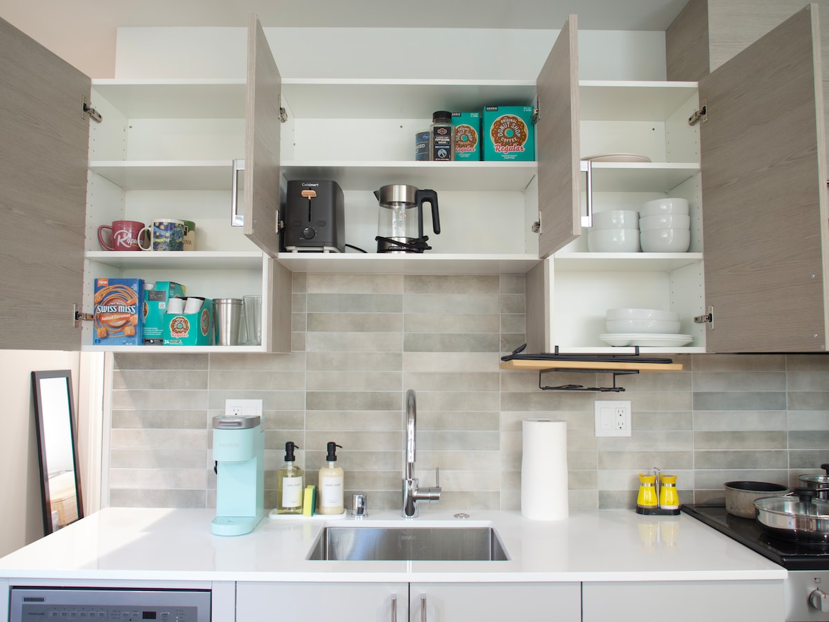 A modern kitchen space features upper cabinets with neatly arranged dishes, coffee supplies, and storage, complemented by a stainless steel sink. A light blue water dispenser and a paper towel roll sit on the white countertop, which is illuminated by natural light.