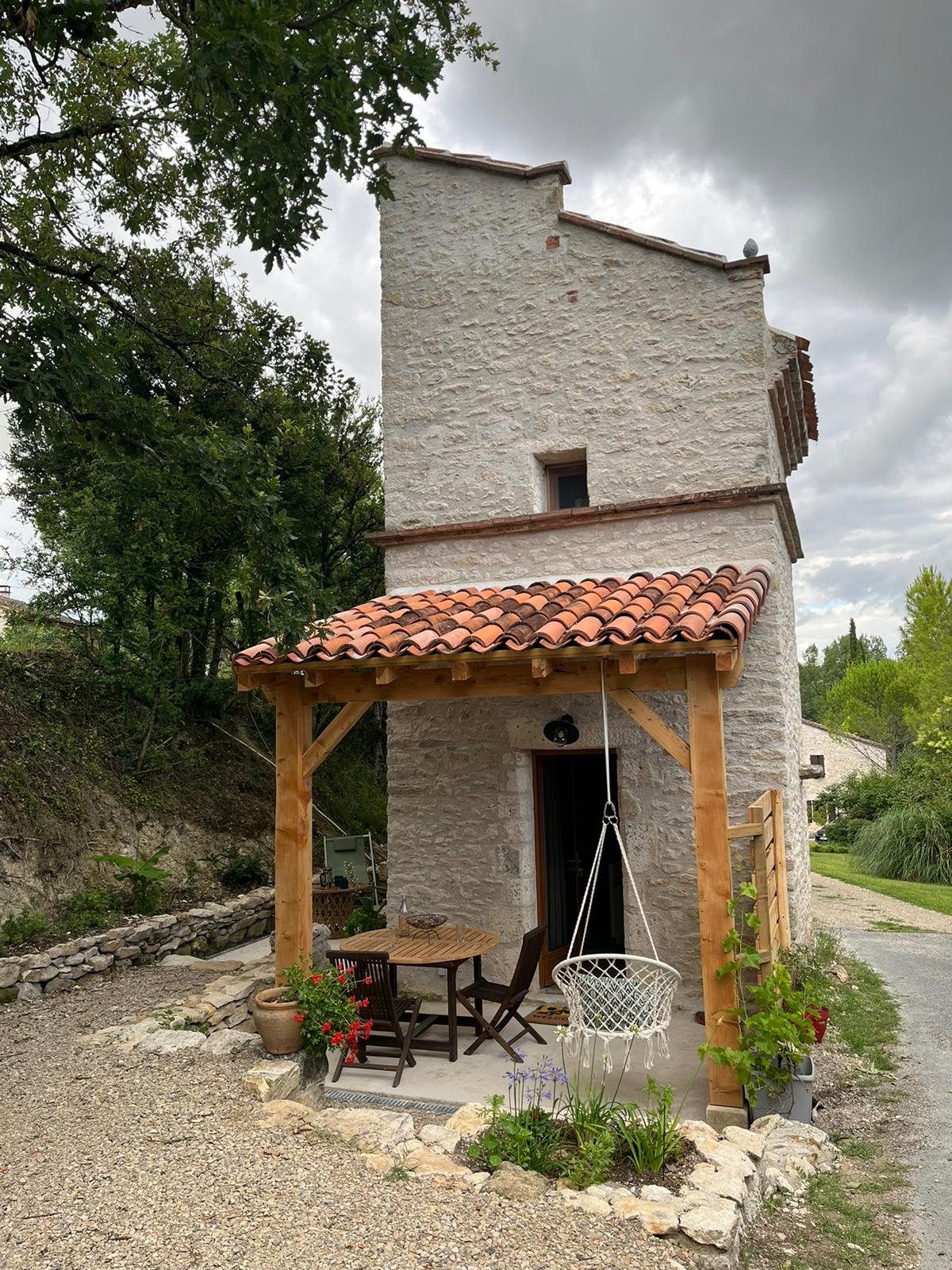 A charming stone building is positioned under a cloudy sky, featuring a terracotta roof and a wooden porch. A small table and chairs are placed on the porch, complemented by potted plants and a hanging chair, inviting relaxation in the outdoor space.