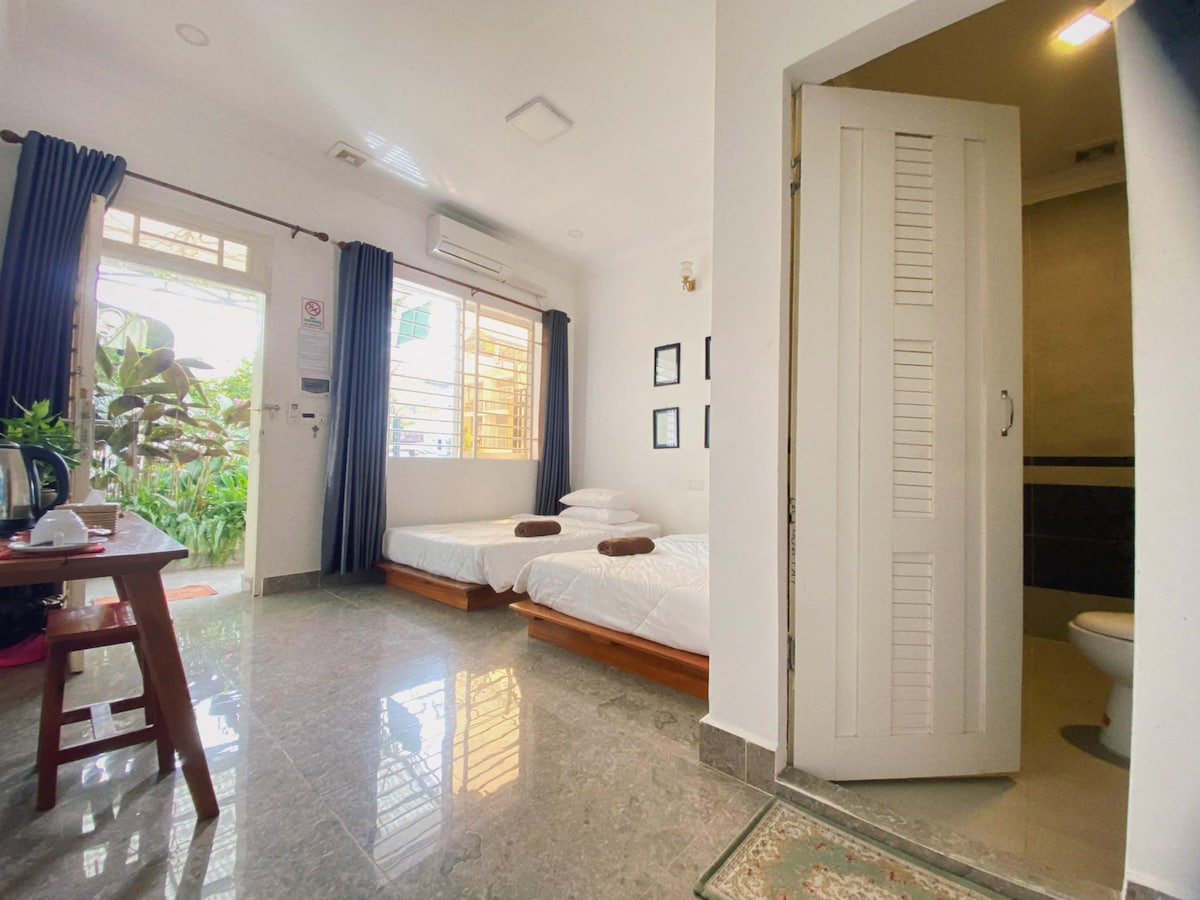 A bright room features two spacious beds with neat linens. Natural light enters through large windows draped with dark curtains, illuminating the polished marble floor. A small table is positioned nearby, and a doorway leads to a well-maintained bathroom with essential amenities.