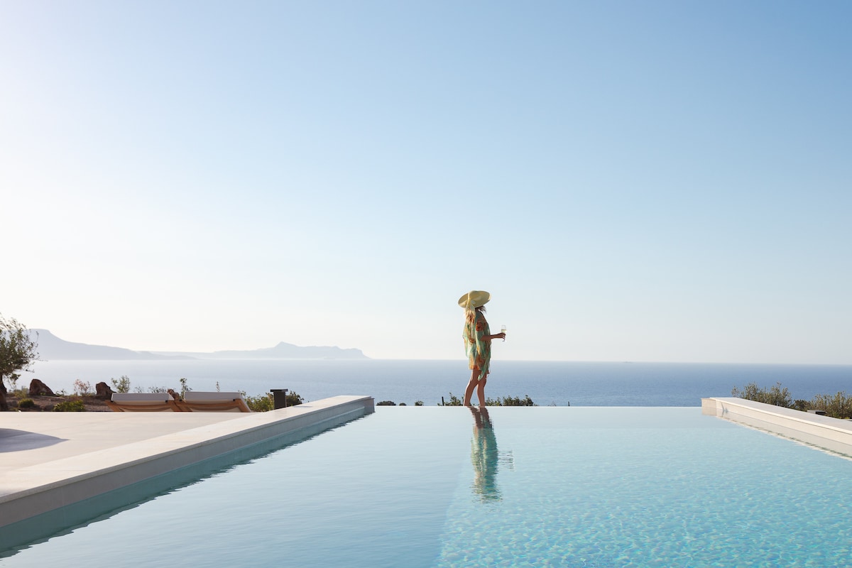 A person stands on the edge of a private infinity pool, gazing out towards the sea, with rolling hills in the distance. The clear blue water of the pool reflects the sky, creating a seamless horizon.