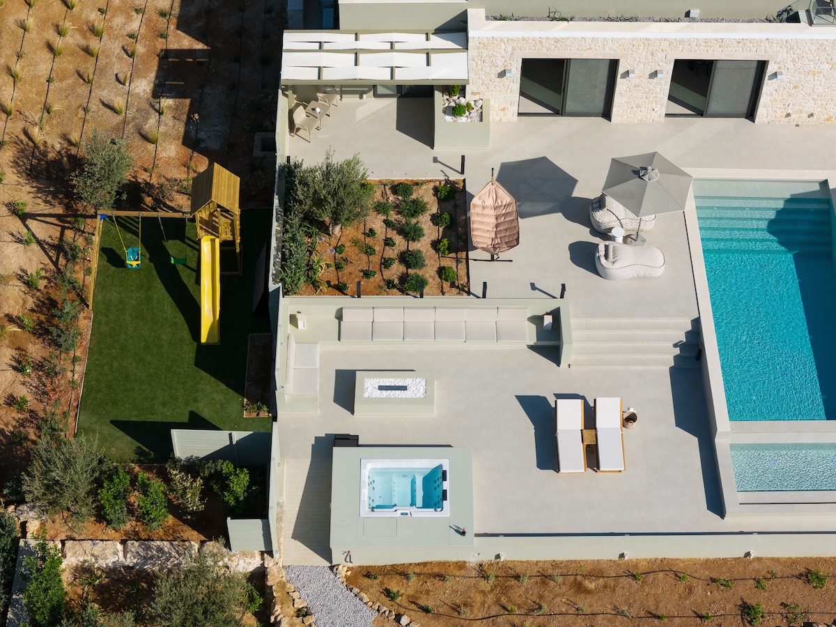 An aerial view of the outdoor area reveals a private eco swimming pool, a six-seater spa whirlpool, and sun loungers arranged for relaxation. A children’s playground featuring a slide is visible, alongside a fire pit and a covered dining space. Olive trees and landscaped areas surround the space.