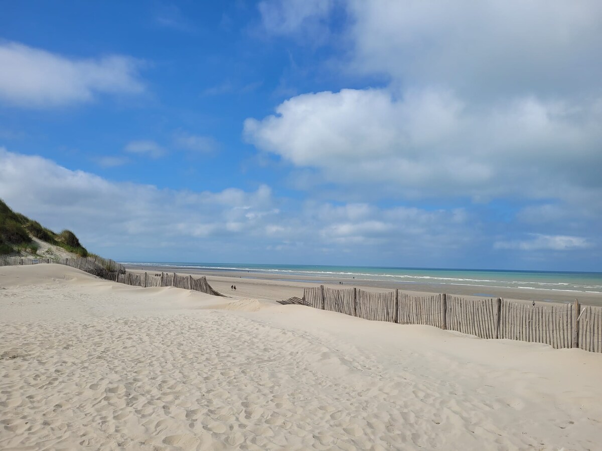 A sandy beach is depicted with gentle dunes leading to a calm sea. Wooden fences define the landscape, and a clear blue sky with scattered clouds complements the tranquil scene. The soft texture of the sand is visible, inviting relaxation by the shore.