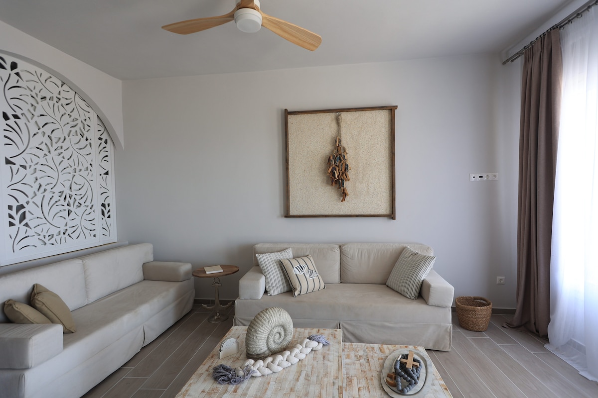 A light-filled living area features two neutral sofas arranged around a wooden coffee table. Decorative accents, such as a shell and a woven basket, add texture. A wall art piece hangs above, and large windows dressed with sheer curtains open to a serene view.