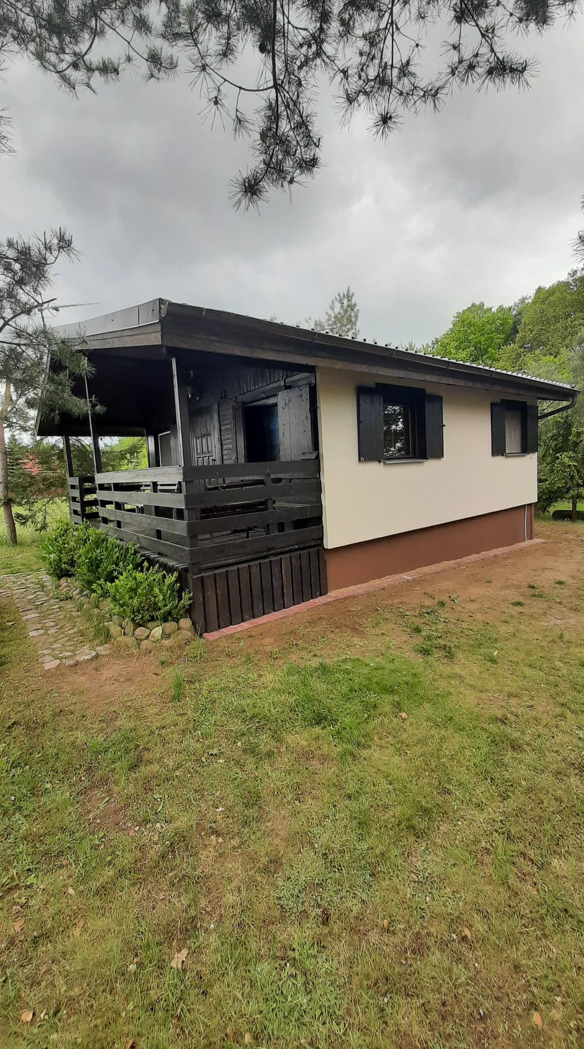 A charming vacation cottage is shown, featuring a dark roof and contrasting light walls. The structure includes numerous windows framed by black shutters. A wooden deck extends from the entrance, surrounded by green grass and trees, offering a peaceful setting.