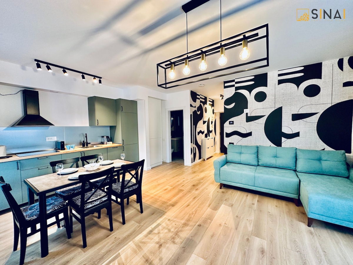 A modern living area features a light blue sofa and a dining table set with four chairs. The kitchen is equipped with sleek appliances, and a distinctive black and white mural adorns one wall. Natural light enhances the space through large windows.