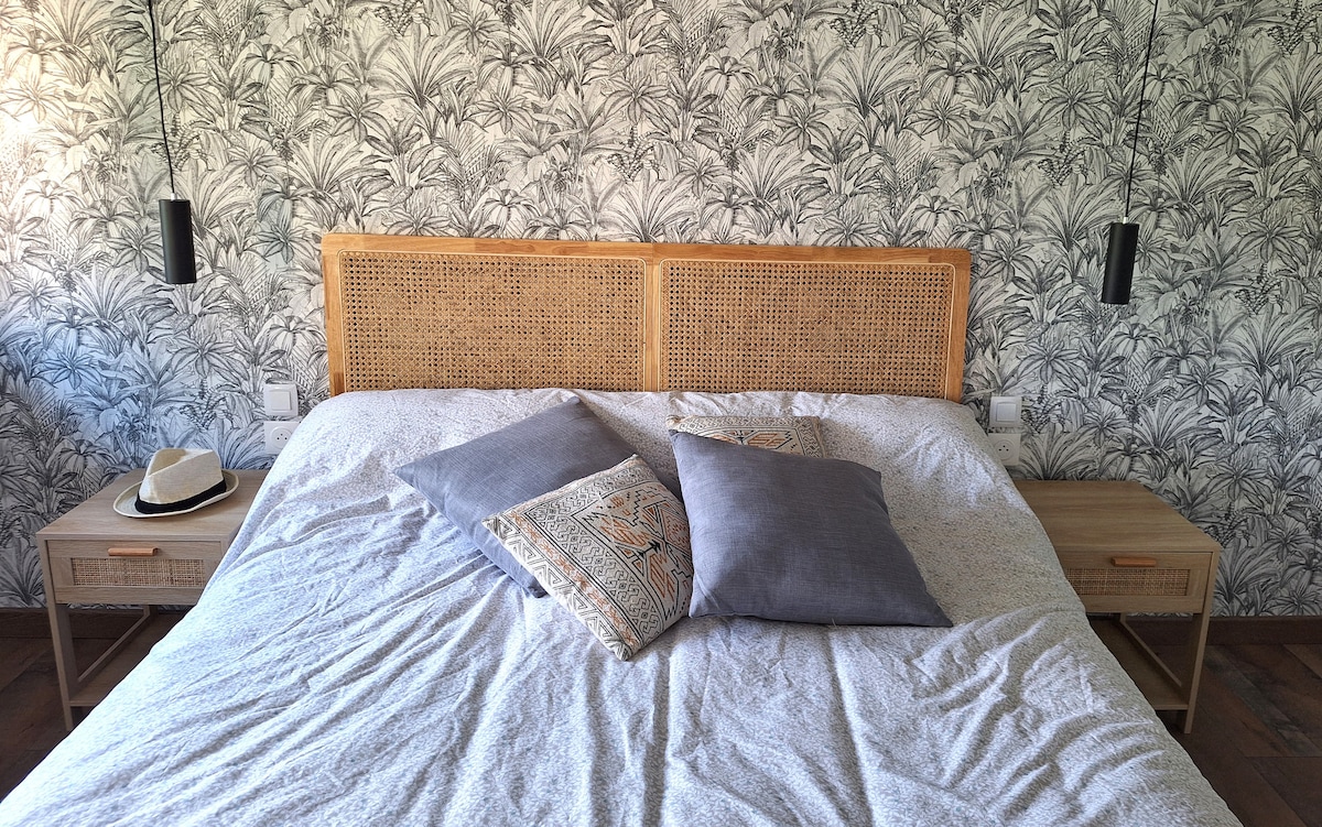 A spacious bedroom features a woven headboard above a large bed dressed in light linens. Two bedside tables with lamps are positioned on either side, complemented by decorative cushions. A patterned wallpaper with a leaf motif enhances the room's ambience.