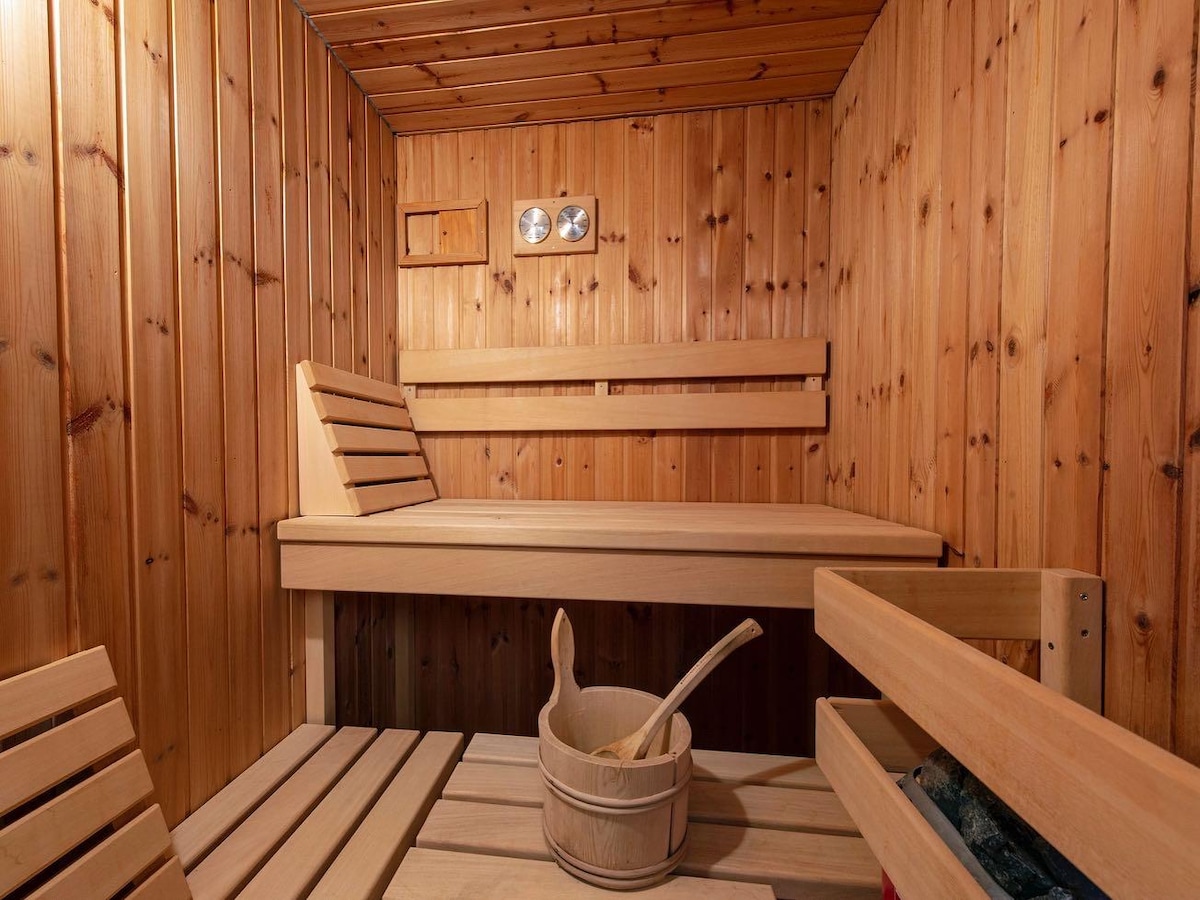 A wooden sauna features slatted benches, offering ample seating. A light-colored wooden bucket and scoop sit on a lower shelf, while wall panels display natural wood grain. Ventilation openings are visible, contributing to the overall design of this calming space.