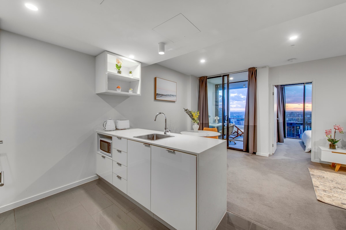A modern kitchen area features sleek white cabinetry and a countertop with a sink. A microwave and kettle are visible on the counter. To the right, large glass doors lead to a balcony with city views, while warm evening light adds to the ambiance.