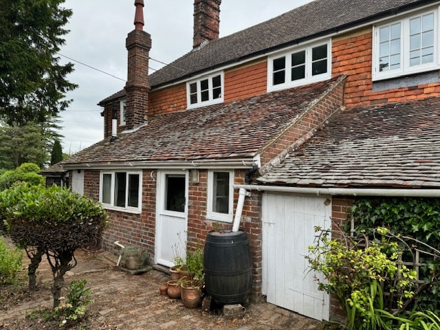 The exterior of an old farmhouse is shown, featuring brick walls and a slate roof. Two chimneys rise from the roof, while windows are positioned above a door. A small patio area with potted plants and greenery surrounds the entrance, enhancing the rustic charm of the property.