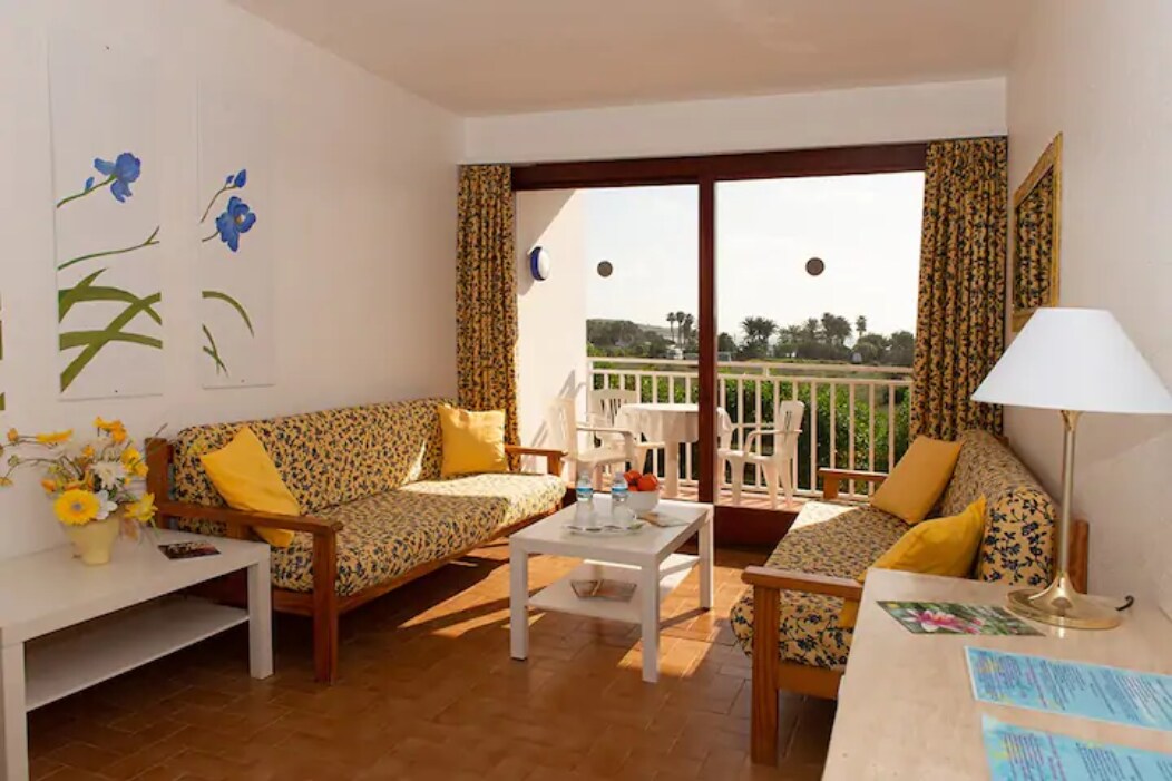 A cozy living area is furnished with two floral-patterned sofas in warm tones, complemented by a simple coffee table. Large glass doors offer views of the outdoor terrace and surrounding greenery, while bright curtains frame the window, enhancing the airy atmosphere.