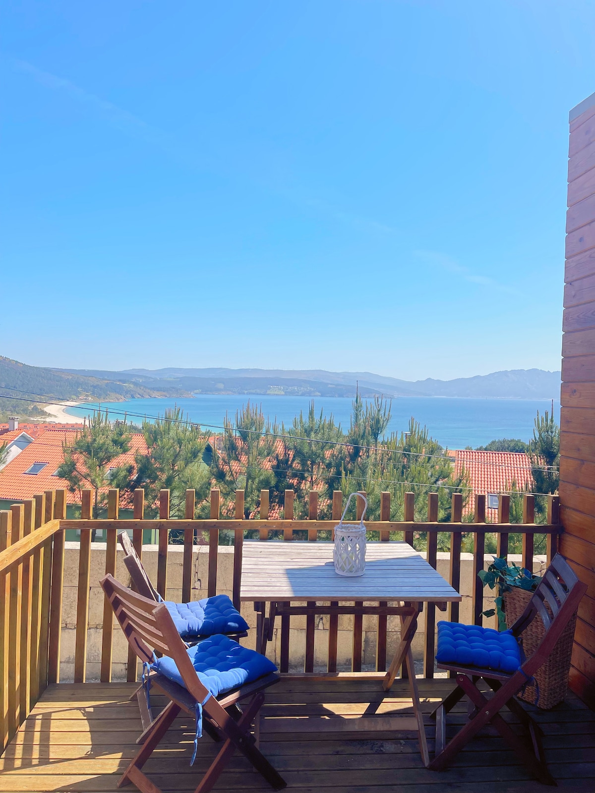 A small wooden balcony overlooks a scenic view of the coastline, with trees and rooftops visible. A round table set with two blue chairs invites relaxation, while a lantern adds a touch of charm. Clear skies enhance the tranquil atmosphere.