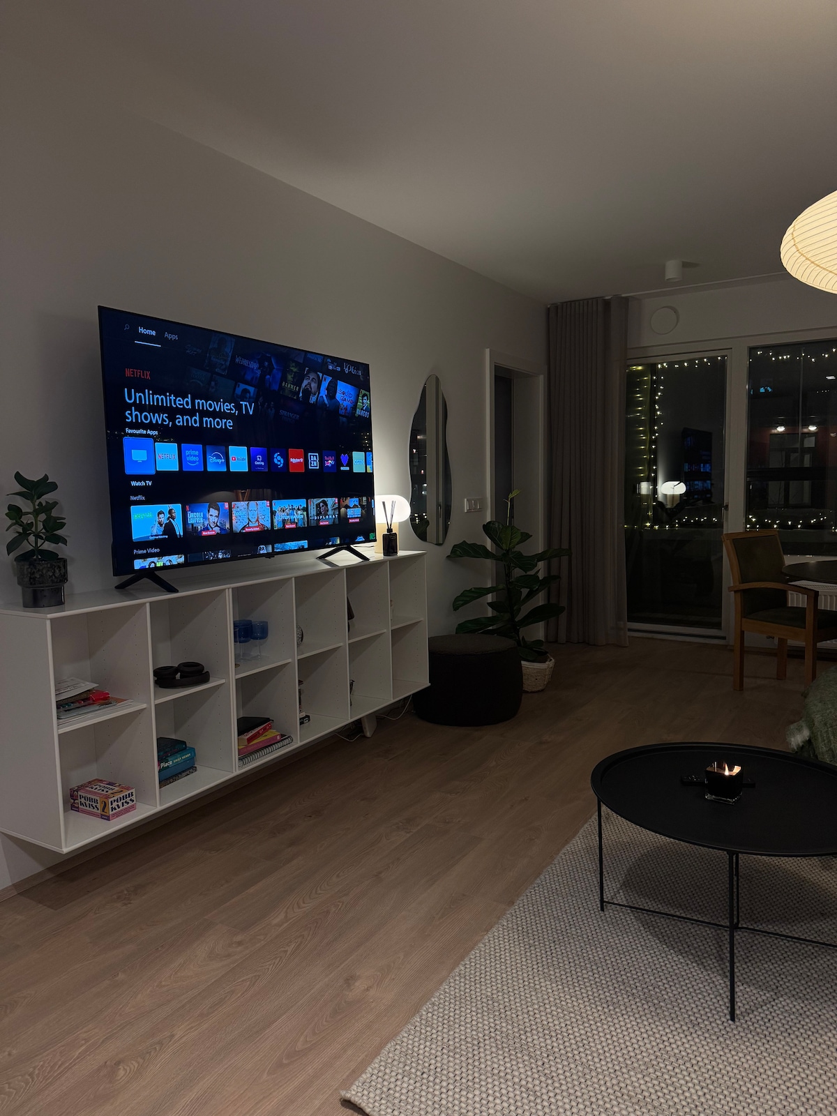 A modern living area features a large flat-screen TV displaying streaming options. A simple white shelving unit holds various items, and a round coffee table is positioned on a textured rug. Natural light is visible through large windows, framed by sheer curtains.