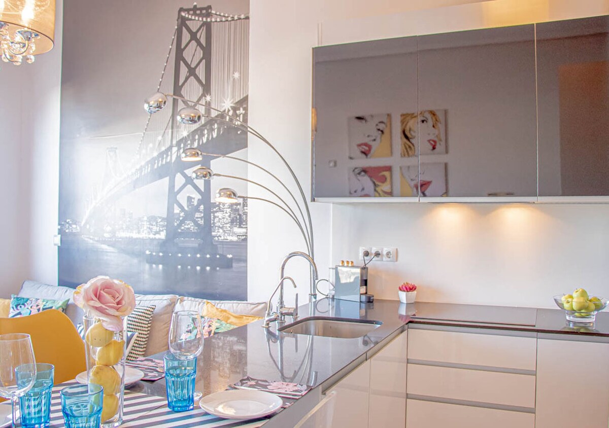 A modern kitchen space is visible, featuring sleek cabinetry and a glossy countertop. Artistic wall decor includes a cityscape and colorful portraits. A dining area is set with blue glasses and decorative items. Soft lighting highlights the design elements, creating a fresh and inviting ambiance.