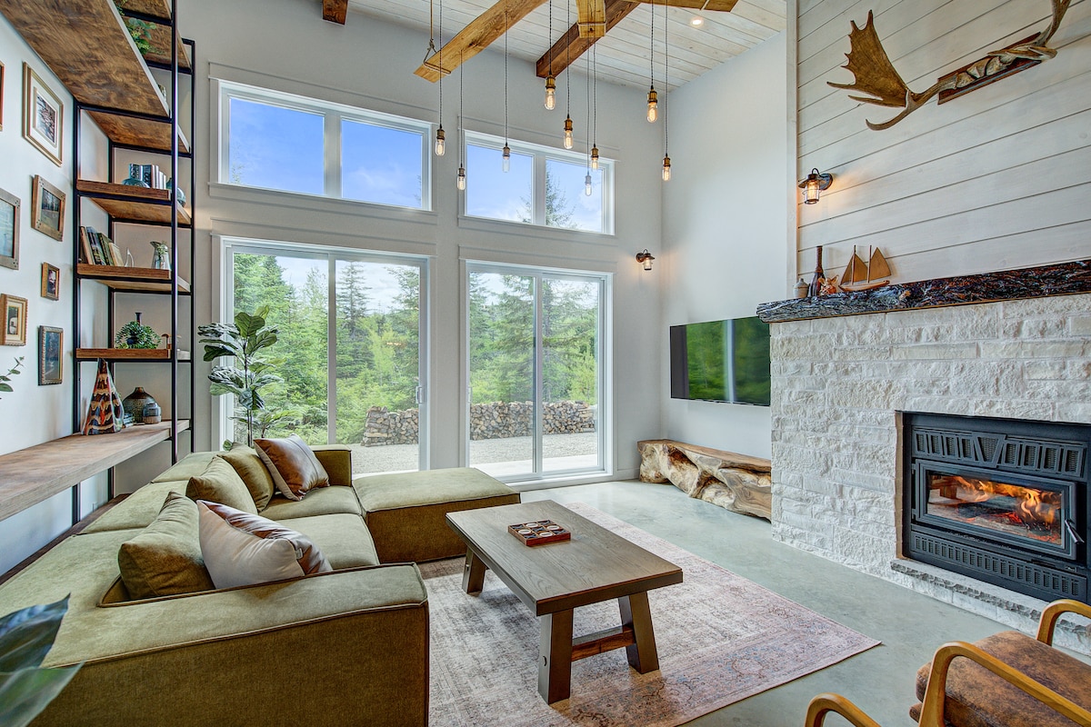 A spacious living area features a large sectional sofa in earthy tones, complemented by a wooden coffee table. Two expansive windows provide a view of the surrounding greenery, while warm light fixtures hang from the high ceiling. A modern fireplace adds warmth to the space.