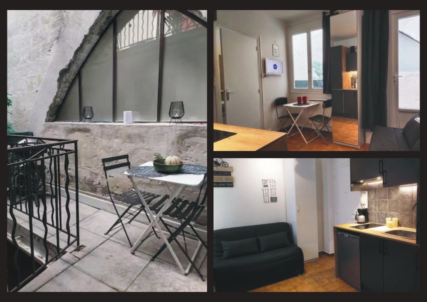 A terrace is equipped with a compact folding table and two chairs, set against a textured stone wall. The adjacent living area features a sleek kitchen counter with modern appliances, a charcoal sofa, and a window allowing natural light to brighten the space.