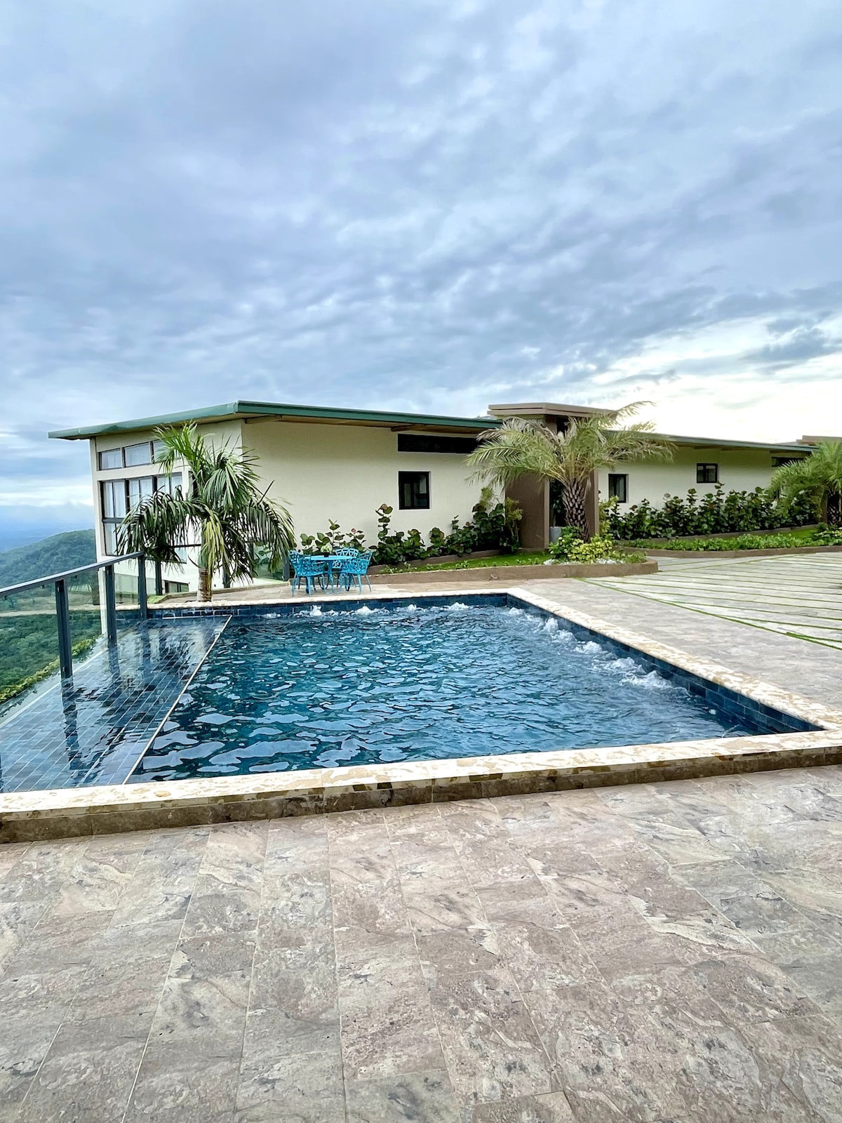 An inviting swimming pool reflects the surrounding landscape, bordered by a sleek glass railing. Lush greenery and palm trees are visible near the modern building, which has glass windows and a spacious deck ideal for relaxation. The sky presents a soft, cloudy backdrop.