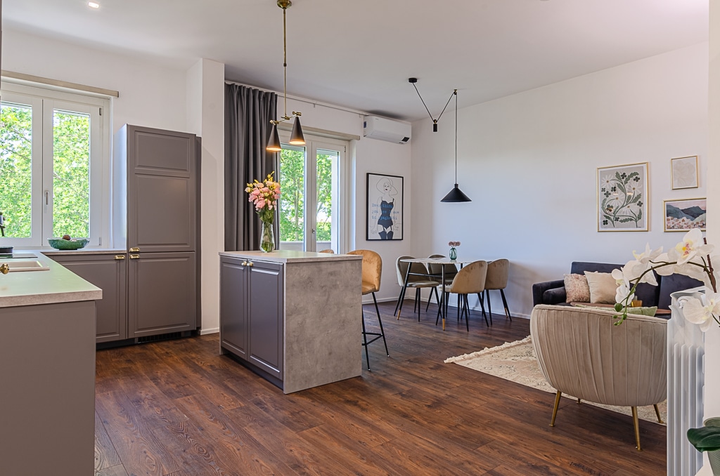 An open-plan living space is showcased, featuring a modern kitchen island with seating and a rustic dining area. Large windows invite natural light, illuminating the sleek furniture. Artwork accents the walls, while a comfortable seating area is positioned in a corner, creating an inviting atmosphere.