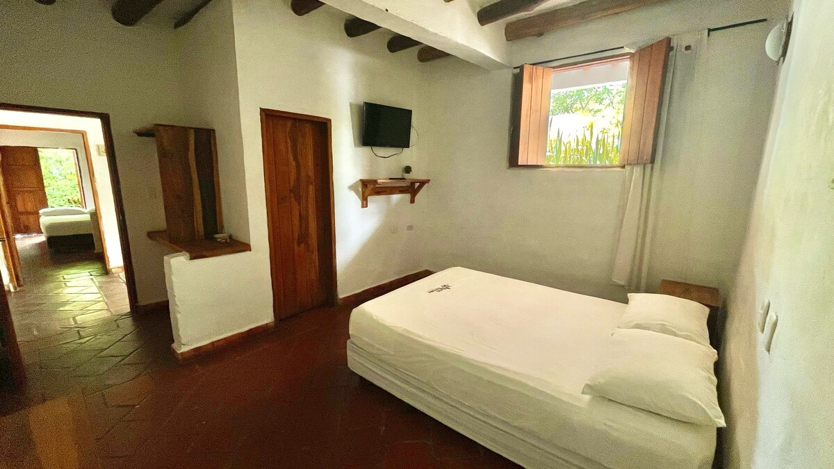 A bright room features a spacious bed with white linens, complemented by wooden shelves and a small television mounted on the wall. Natural light filters in through a window framed by light curtains, shedding warmth on the terracotta tile floor.