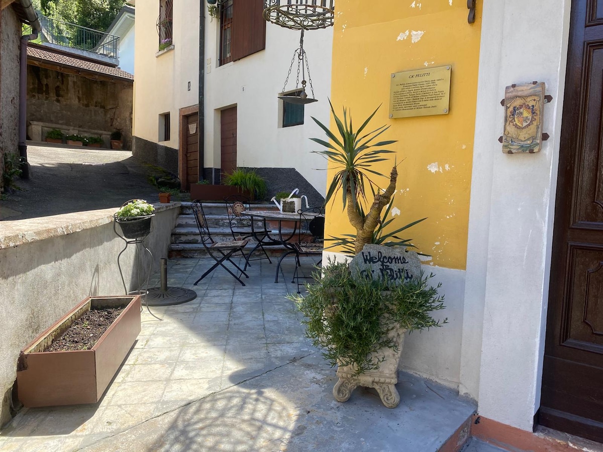 An outdoor patio area features a decorative planter with a green palm and a welcoming sign. A small dining table is accompanied by chairs, and a set of stone steps leads towards a nearby entrance, suggesting a serene and inviting entryway.