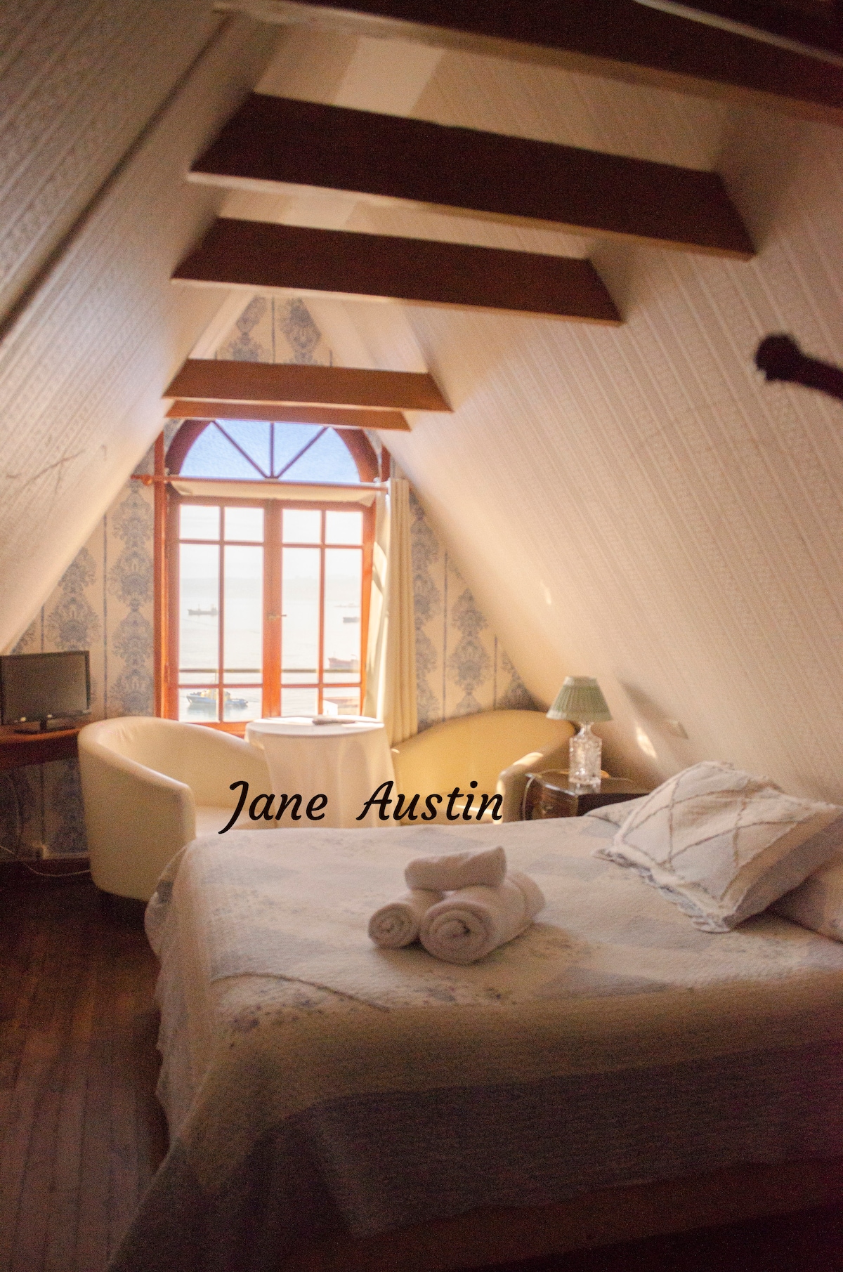 A serene room features a spacious bed adorned with a light-colored blanket. A large window captures ample sunlight, framed by blue wallpaper and exposed wooden beams. A cozy seating area includes a curved chair and a small table, providing a relaxing corner.