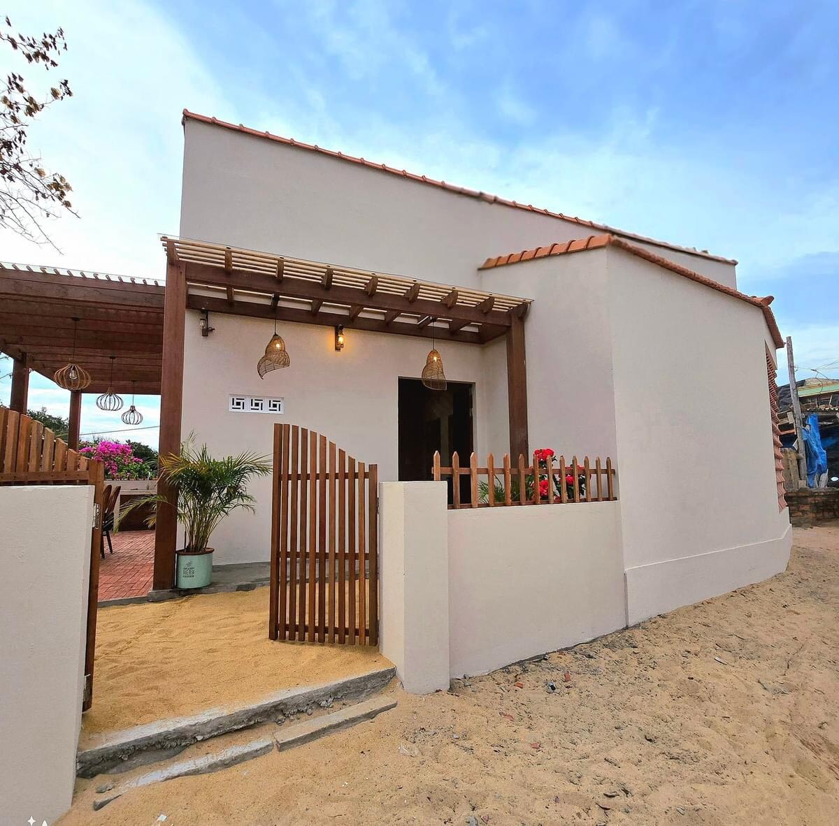A modern house is displayed with a smooth exterior and a terracotta roof. A wooden fence borders the entrance, while decorative lanterns are suspended above. The sandy surroundings contribute to the coastal aesthetic, enhancing accessibility to the nearby beach.
