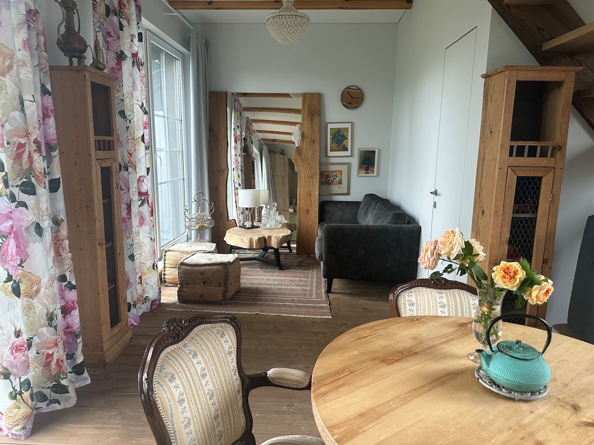 A quaint living area features a comfortable dark sofa, a round wooden dining table, and elegant floral curtains. A large mirror reflects the space, enhancing the light. Decorative pieces, including a turquoise kettle on the table and a vase of fresh flowers, add charm.