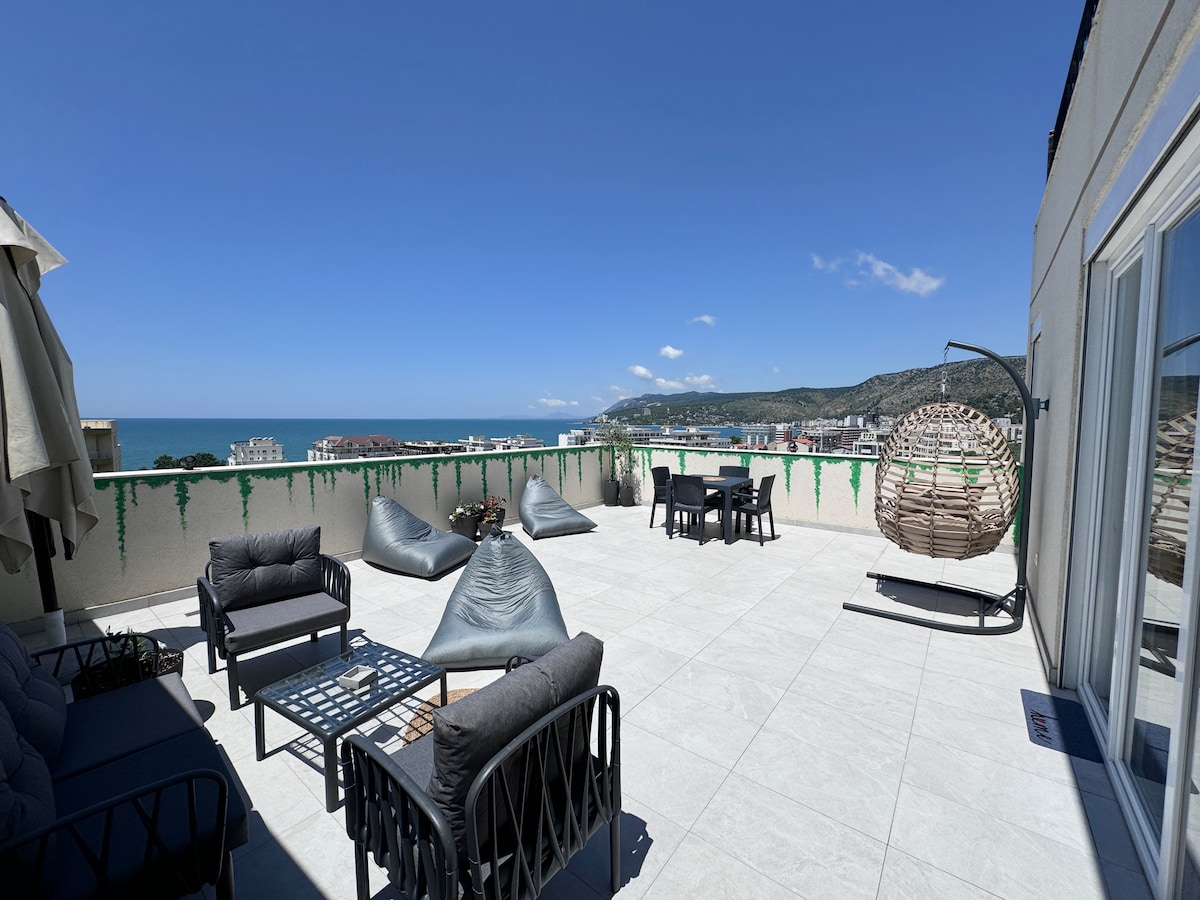 A spacious rooftop terrace features comfortable seating arrangements, including lounge chairs and a hanging chair. The area is enhanced by a panoramic view of the coastline and mountains. Sunlight reflects off the tiled floor, creating a bright and inviting outdoor space.