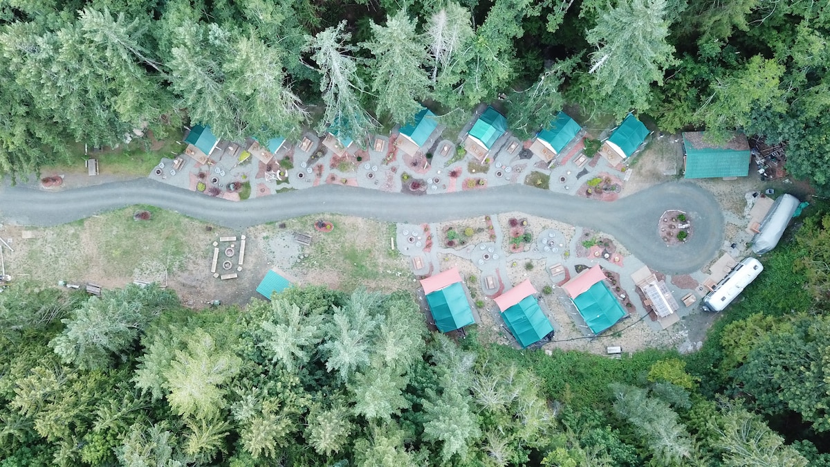 An aerial view captures the glamping village nestled among lush green trees. A winding path connects individual safari tents, each featuring colorful roofs. The landscape includes flower beds and seating areas, set against the serene backdrop of dense forest, promoting a peaceful retreat.