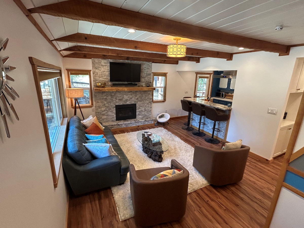 A spacious living area features wooden beams overhead and a stone fireplace as the focal point. Comfortable seating is provided by a leather sofa and two armchairs, arranged around a decorative coffee table placed on a soft rug. Natural light enters from large windows.