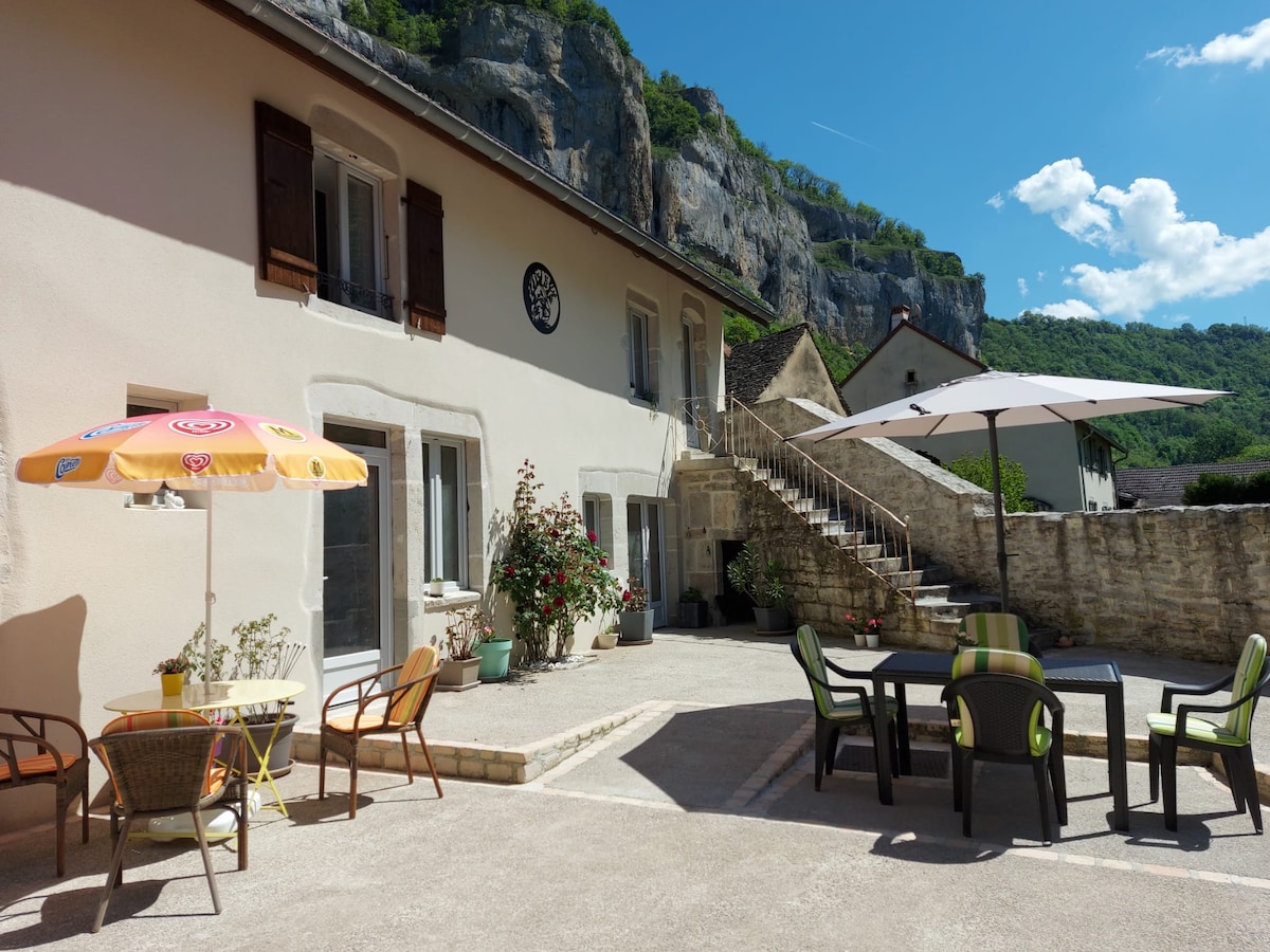 A shared outdoor space features a spacious patio with several seating arrangements, including a round table with chairs and a colorful umbrella. Surrounding greenery and a cliffside can be seen in the background, providing a peaceful setting for relaxation.